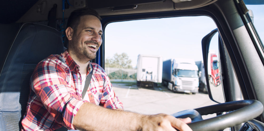 A smiling semi truck driver in a flannel shirt steers around a parking lot.